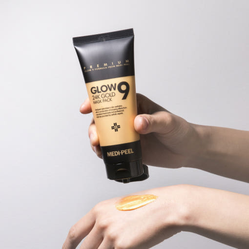 Hand holding a tube of 'Glow 24K Gold Mask Pack' above a wrist with a small amount of the mask applied.
